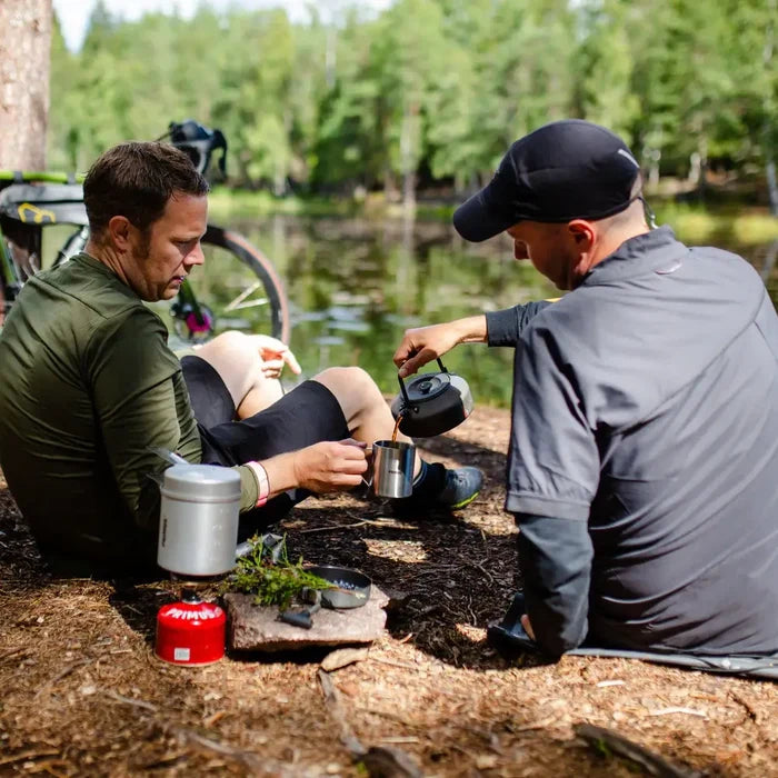 Two men camping by a lake, one pouring tea from a Primus 900ml Litech kettle into a cup.