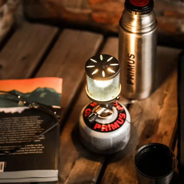 Primus branded thermos and stainless steel Micron lamp on a wooden surface with books in the background