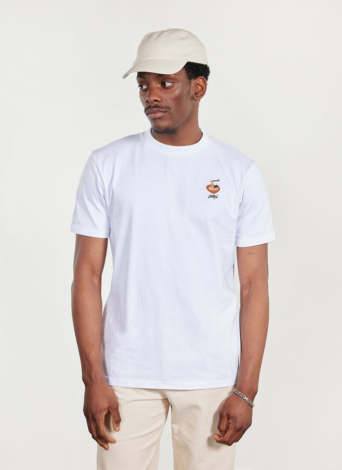 Model wears Percival Ramen T-Shirt in white organic cotton, featuring a ramen bowl embroidery, with a cream cap and trousers.