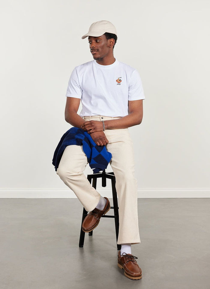 A model sits wearing the Percival white organic cotton t-shirt with ramen embroidery, styled with cream trousers and a cap