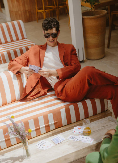 A man in a Percival rust linen suit and sunglasses smiles while playing cards on a striped outdoor sofa.