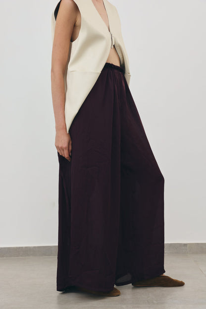 A model is shown from the side wearing a cream sleeveless vest and dark plum wide-leg trousers, paired with brown shoes, against a plain light gray studio background.