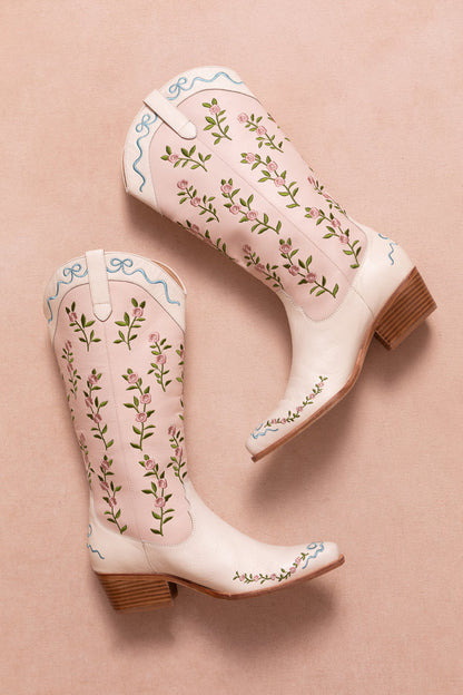 The Rosemary Cowgirl Boots by Ivy City Co are light pink leather boots with an embroidered floral pattern and blue trim, displayed on a soft pink background, both boots lying on their sides with heels angled outward.