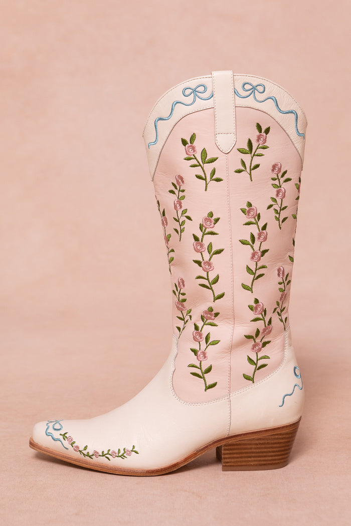 The Rosemary Cowgirl Boots by Ivy City Co are light pink with embroidered green stems, pink flowers, blue trim at the top and toe, and a low wooden heel, shown on a pale pink background.