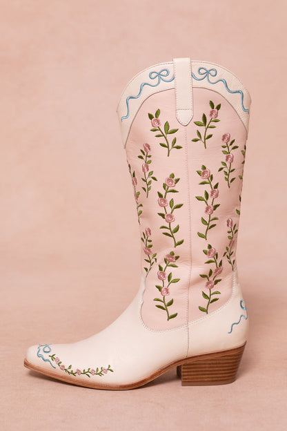 The Rosemary Cowgirl Boots by Ivy City Co are light pink with embroidered green stems, pink flowers, blue trim at the top and toe, and a low wooden heel, shown on a pale pink background.