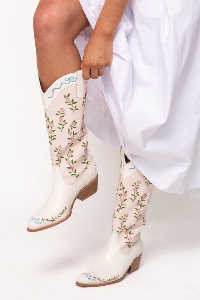 A person in a white dress adjusts their cream Ivy City Co Rosemary Cowgirl Boots, featuring embroidered floral patterns and light blue accents.