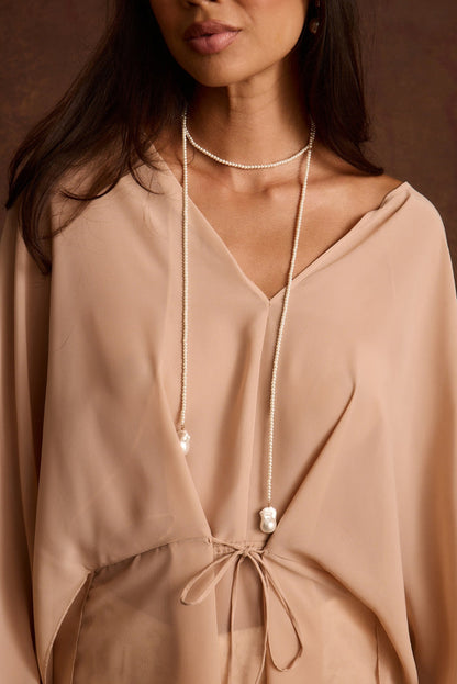 Long beaded pearl necklace with a baroque pearl drop pendant, worn over a tied blush blouse.