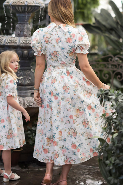 A woman and a young girl wear matching Ivy City Co Sophia Dresses in Countryside Peony Blossom near a garden fountain. The woman faces away, holding the girl's hand, as the girl in her vintage-inspired dress looks at the camera.