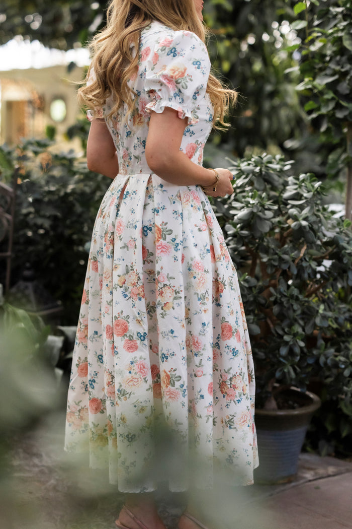 A woman with long wavy hair stands among green plants, her back to the camera, wearing Ivy City Co’s vintage-inspired Sophia Dress in Countryside Peony Blossom.