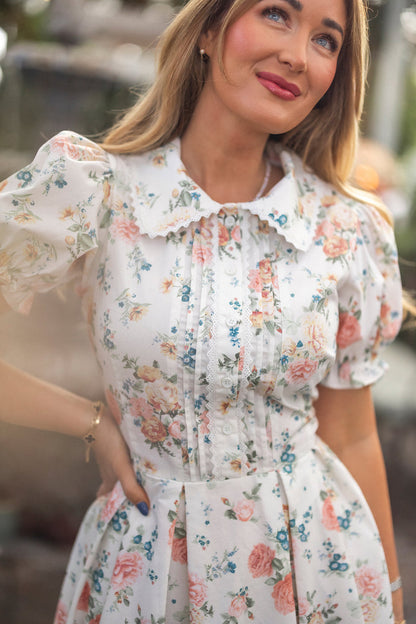 A woman wearing Ivy City Co’s Sophia Dress in Countryside Peony Blossom stands outdoors, smiling softly with one hand on her hip. The vintage-inspired dress features a ruffled collar, puffed sleeves, and vertical pleats on the bodice.