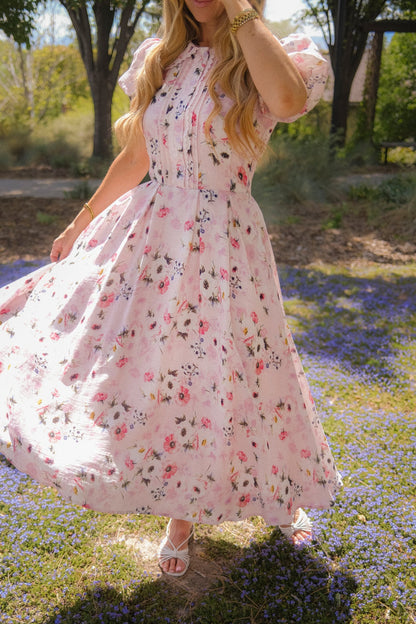 A person with long wavy hair, wearing the Ivy City Co Sophia Dress in Roselyn Pink and white sandals, twirls outdoors on a field of purple flowers, holding the hem. Their face is not visible.