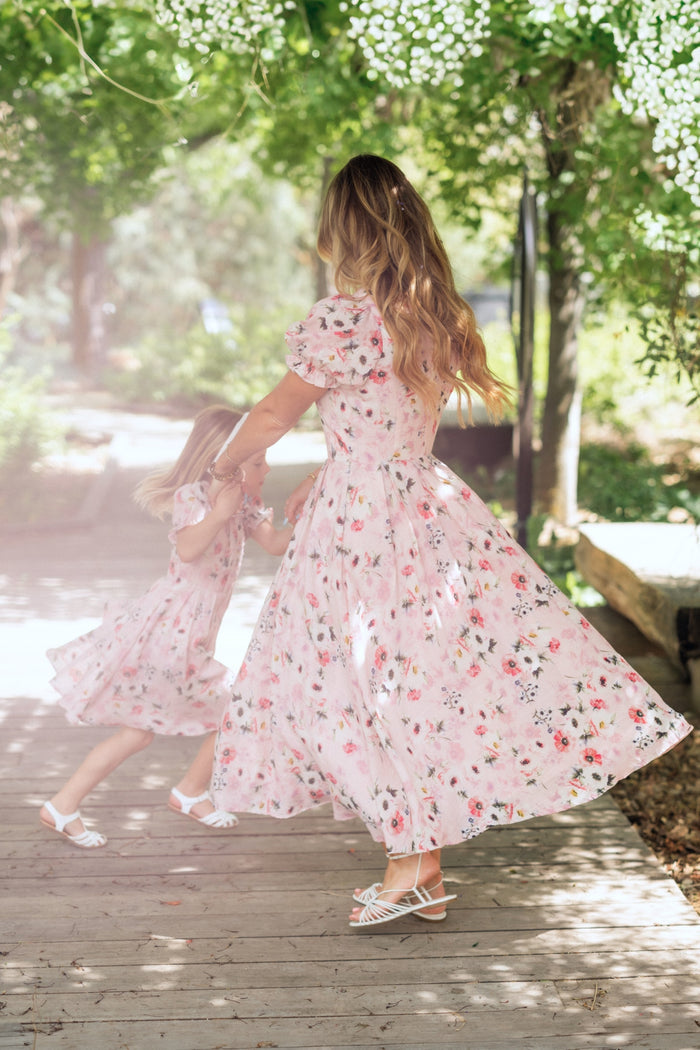 A woman and a young girl twirl together outdoors in matching Ivy City Co Sophia Dresses in Roselyn Pink, surrounded by greenery and sunlight on a wooden path.