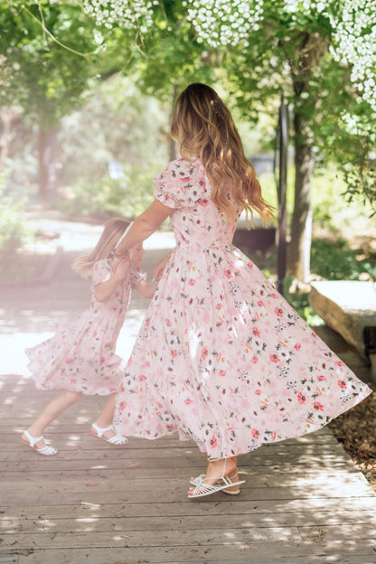 A woman and a young girl twirl together outdoors in matching Ivy City Co Sophia Dresses in Roselyn Pink, surrounded by greenery and sunlight on a wooden path.