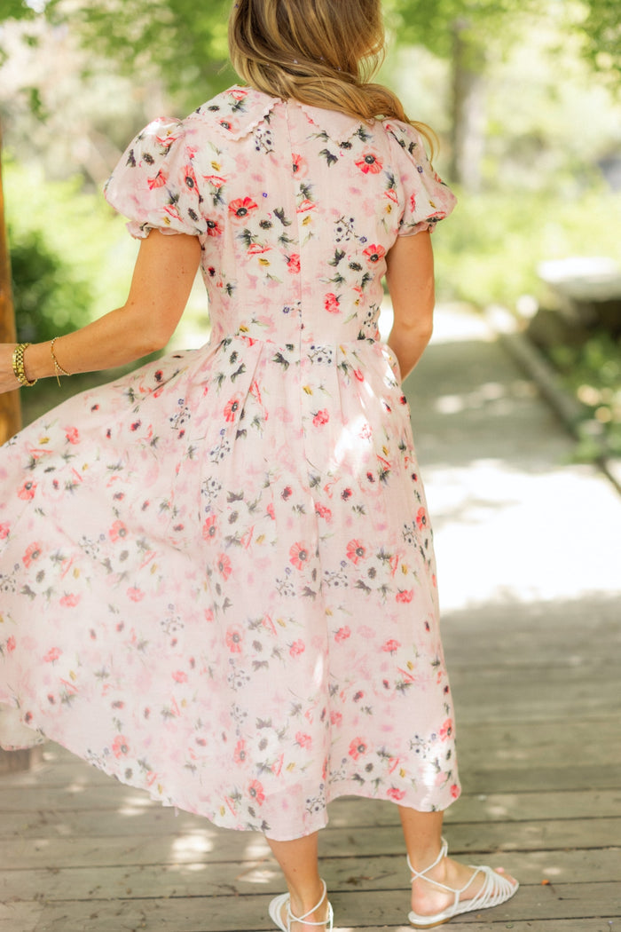 A woman with long hair wears the Ivy City Co Sophia Dress in Roselyn Pink with floral patterns and short puff sleeves, paired with white sandals, standing on a wooden pathway outdoors amid greenery and sunlight.
