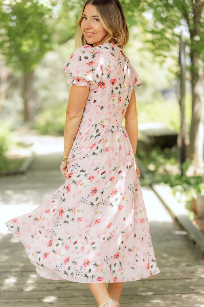 A woman wearing the Ivy City Co Sophia Dress in Roselyn Pink stands on a wooden path outdoors, turning and smiling at the camera. Sunlight filters through lush green trees, highlighting her floral cotton-linen dress.