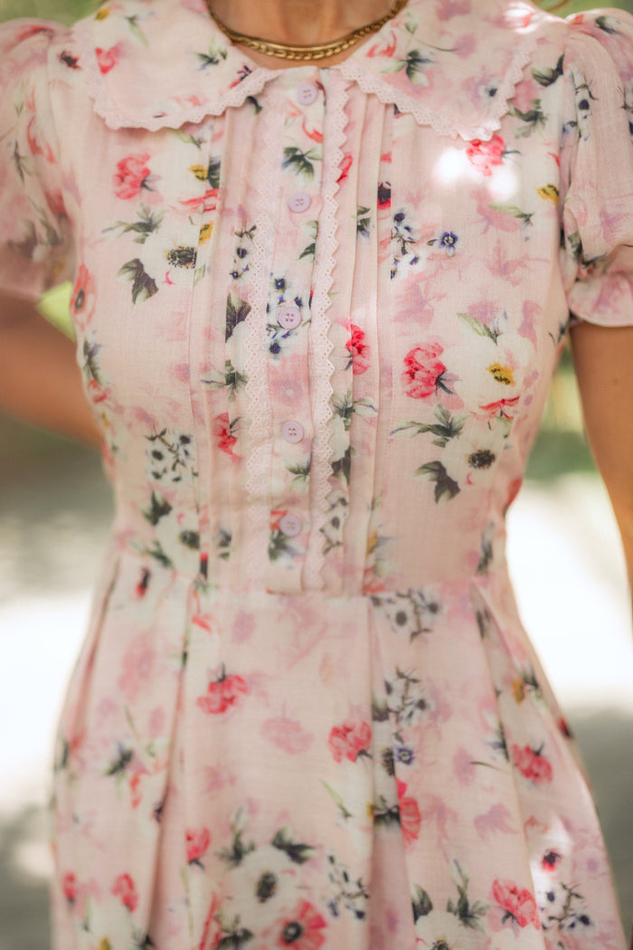 A close-up of a person wearing the Ivy City Co Sophia Dress in Roselyn Pink, a vintage-inspired midi dress with buttons, pleats, scalloped collar, flower pattern, puffed short sleeves, and styled with a gold necklace.