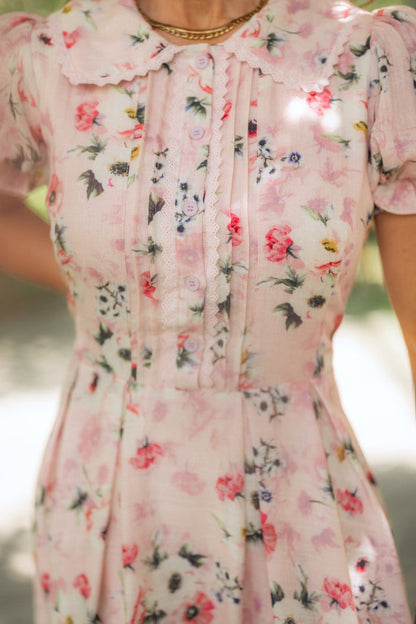 A close-up of a person wearing the Ivy City Co Sophia Dress in Roselyn Pink, a vintage-inspired midi dress with buttons, pleats, scalloped collar, flower pattern, puffed short sleeves, and styled with a gold necklace.