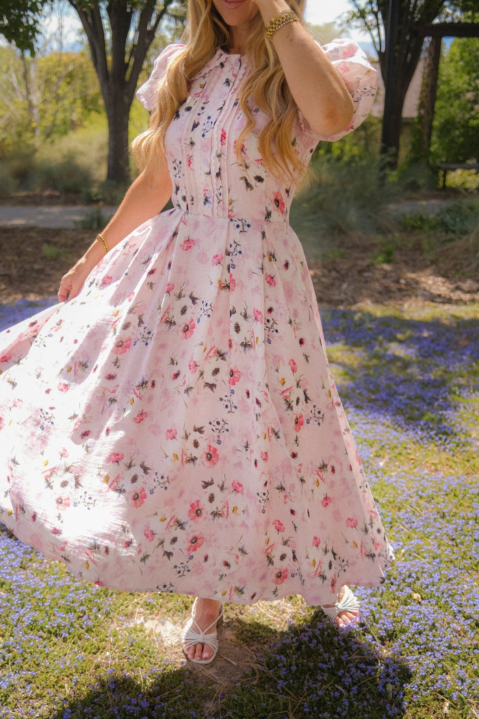 A woman wearing the Ivy City Co Sophia Dress in Roselyn Pink twirls outdoors on a sunny day, standing on a patch of purple flowers with trees in the background. Her face is partially obscured by her hand and hair.