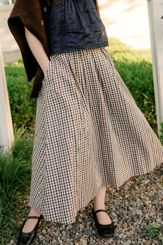 Standing on gravel, the person is wearing the Ivy City Co Sophia Skirt in Chestnut Gingham with pockets, paired with a black top, brown shawl, and black shoes, hands tucked into the skirt’s pockets. The background features greenery and a white structure.