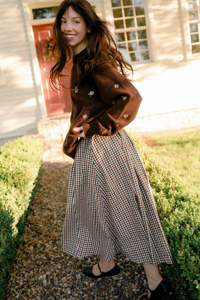 A woman with long brown hair smiles as she walks outside on a gravel path, dressed in a brown sweater adorned with small flowers and the Ivy City Co Sophia Skirt in Chestnut Gingham—a charming cotton skirt with pockets. She is near bushes and a light-colored house with large windows.