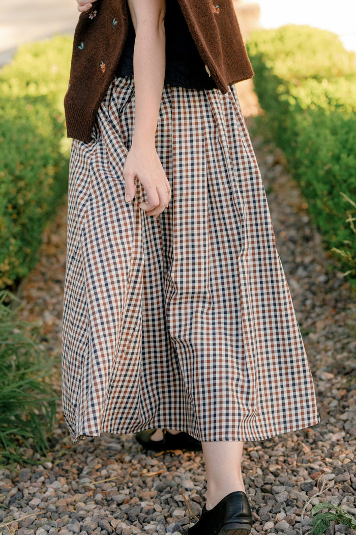 A person wearing a brown vest and the Ivy City Co Sophia Skirt in Chestnut Gingham, a long plaid cotton skirt with pockets, walks on a gravel path between green bushes, their hand relaxed by their side.