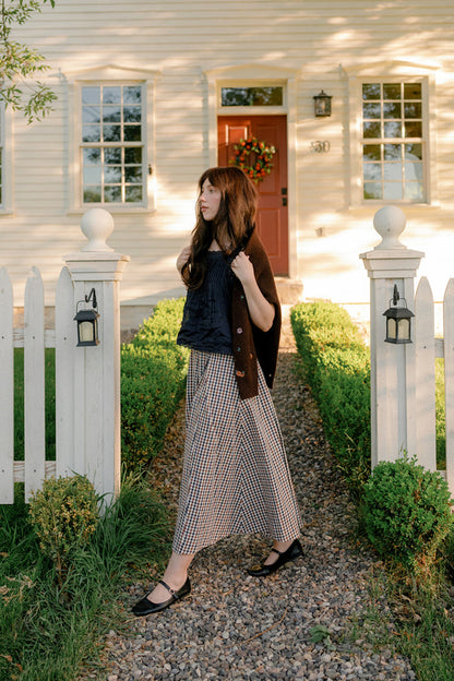 A woman wearing the Ivy City Co Sophia Skirt in Chestnut Gingham with pockets, paired with a black top and brown cardigan, stands on a stone path in front of a white house with a red door and white picket fence on a sunny day.