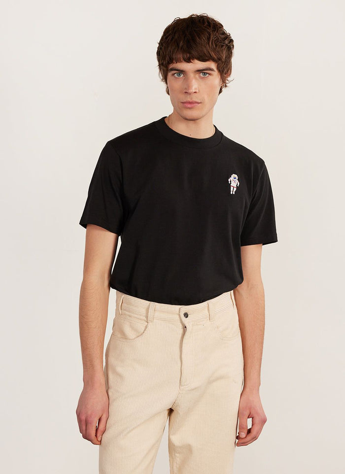 A model wears the black Percival Spaceman embroidered T-shirt with cream corduroy trousers.
