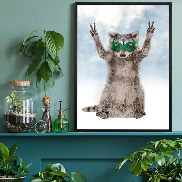 Watercolor raccoon superhero art print wearing a green mask, framed in black on a teal wall with indoor plants.