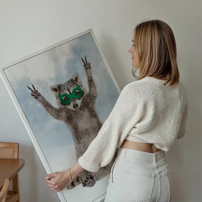 Woman holding a large framed watercolor print of a raccoon superhero wearing a green mask against a cloudy sky.