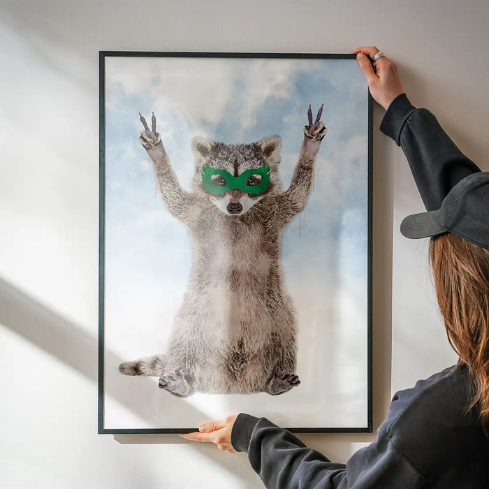 Watercolor raccoon superhero poster with a green mask, making peace signs against a cloudy sky in a black frame.
