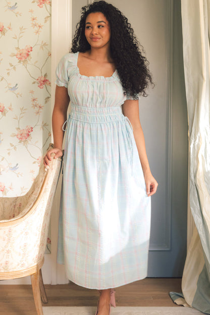 A woman with long curly hair wears the Ivy City Co Thalia Dress in Pastel Plaid, standing indoors beside a floral chair and floral wallpaper, near a blue-gray door as soft light filters through the curtains.