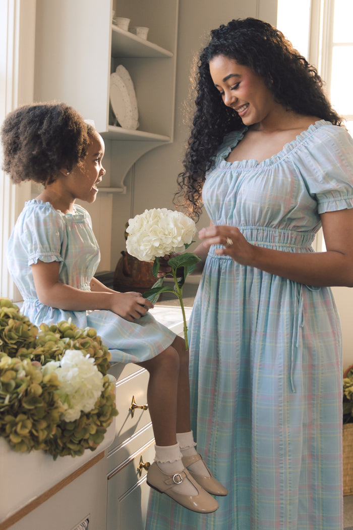 A woman and a young girl share a smile in a sunlit kitchen, both wearing the Ivy City Co Thalia Dress in Pastel Plaid. The girl sits on the counter with a white hydrangea, while the woman stands beside her holding another flower.