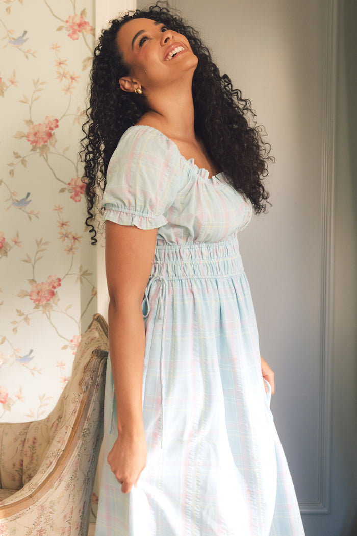 A woman with curly hair, dressed in the Thalia Dress in Pastel Plaid by Ivy City Co, stands indoors smiling under soft sunlight. Floral wallpaper and a vintage chair complete this charming spring-inspired scene.