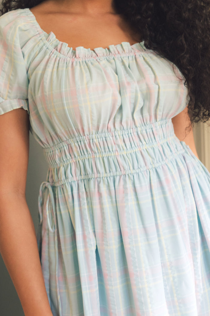 A person wears the Ivy City Co Thalia Dress in Pastel Plaid, featuring short sleeves, a ruched elastic waist, and a ruffled neckline. The light blue dress has pink and yellow stripes—a perfect spring look with visible curly hair.