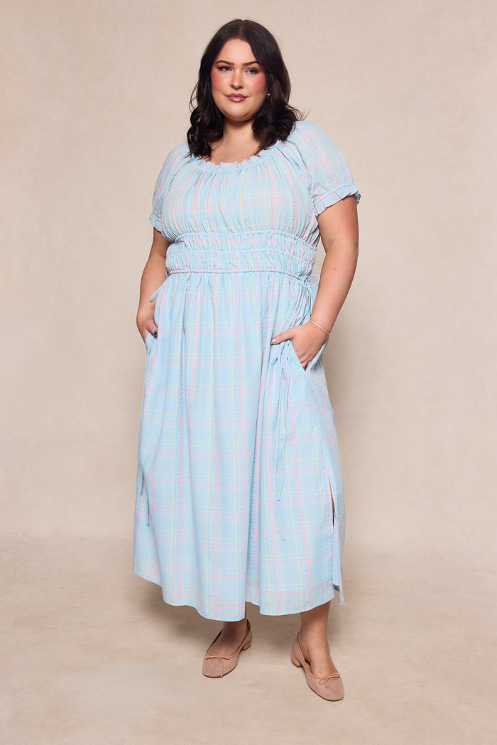 A woman with medium-length dark hair wears the Ivy City Co Thalia Dress in Pastel Plaid, featuring pockets, and nude flats. She stands against a neutral background with her hands in the dress pockets, looking at the camera.