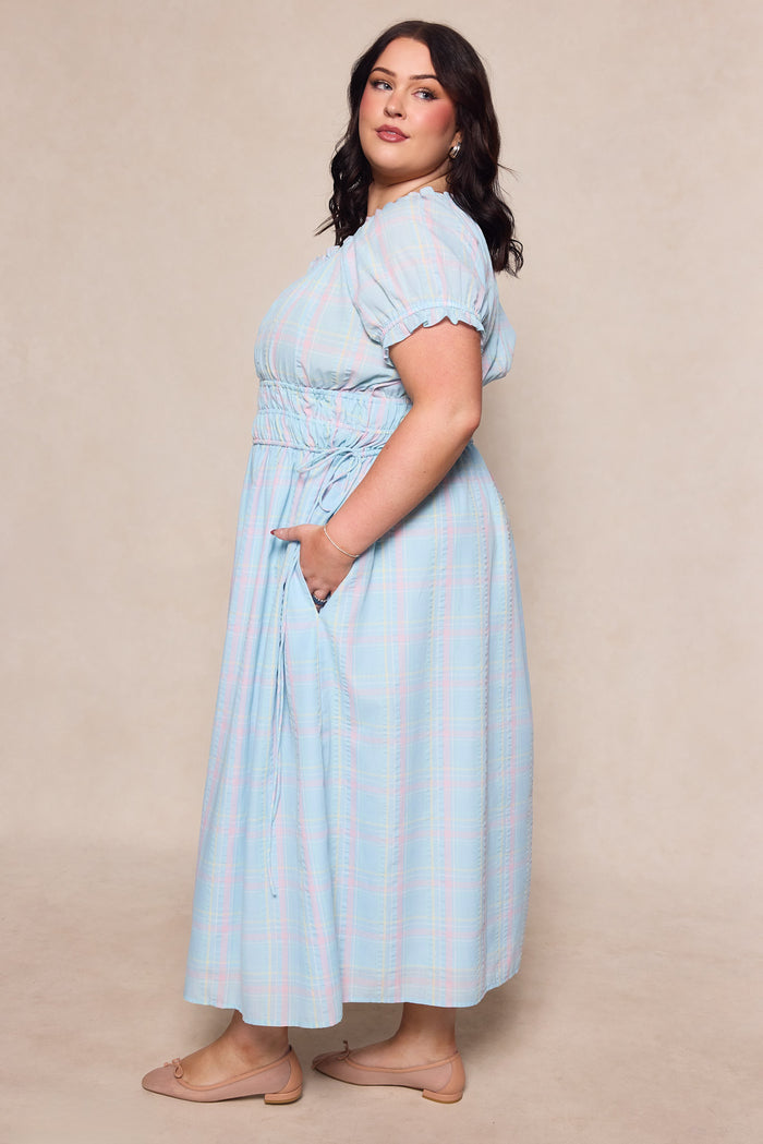 A woman with medium-length dark hair models the Ivy City Co Thalia Dress in Pastel Plaid—a maternity-friendly style with a cinched waist and pockets—standing in profile, hand in pocket, wearing nude flats against a neutral background.