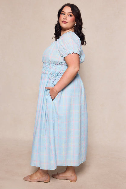 A woman with medium-length dark hair models the Ivy City Co Thalia Dress in Pastel Plaid—a maternity-friendly style with a cinched waist and pockets—standing in profile, hand in pocket, wearing nude flats against a neutral background.