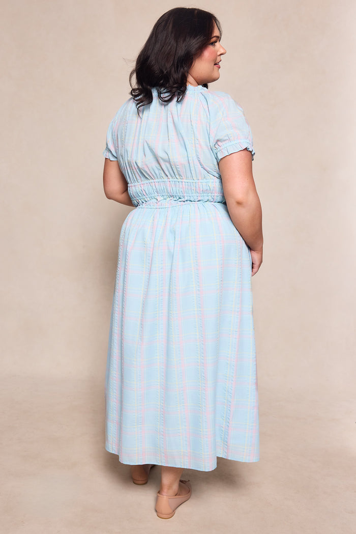 A woman with dark shoulder-length hair wears the Ivy City Co Thalia Dress in Pastel Plaid—a light blue, short-sleeved, ankle-length dress with a gathered waist—and tan flats, standing on a neutral background.