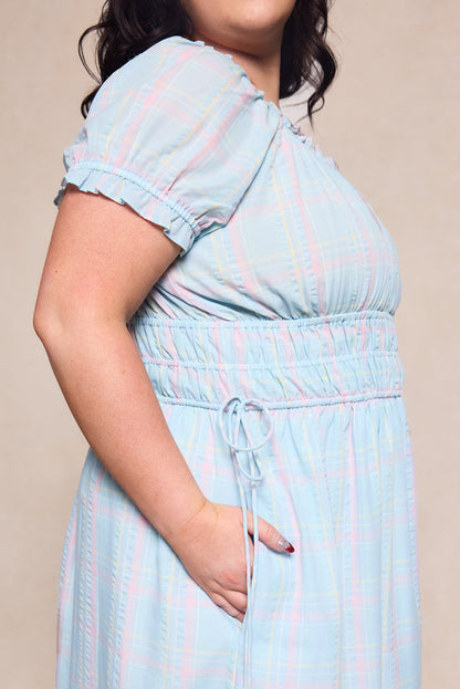 A woman models the Thalia Dress in Pastel Plaid by Ivy City Co, a maternity-friendly spring dress with short puff sleeves, elastic waist, and pockets—shown from shoulders to mid-thigh for a comfy and stylish look.