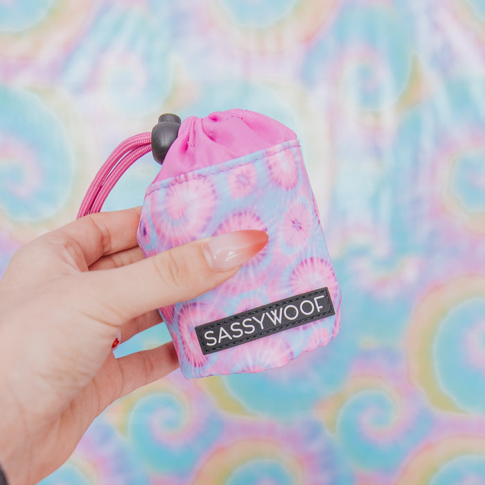 A hand with long, manicured nails holds the Sassy Woof Treat Pouch - Purple Haze, a colorful drawstring accessory from the dog collection, in front of a pastel tie-dye background—ideal for pairing with matching harnesses.