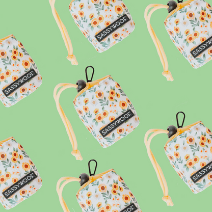 The Sassy Woof “Treat Pouch - Sunflower Dreams” features a white floral design with orange flowers, green leaves, and yellow straps on a light green background—stylish and perfect for pet owners.