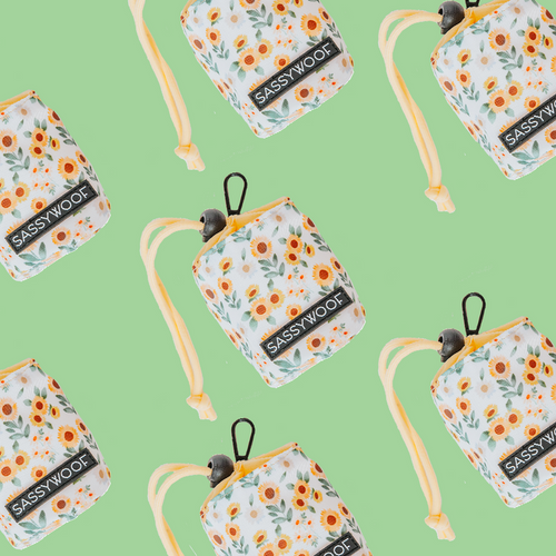 The Sassy Woof “Treat Pouch - Sunflower Dreams” features a white floral design with orange flowers, green leaves, and yellow straps on a light green background—stylish and perfect for pet owners.