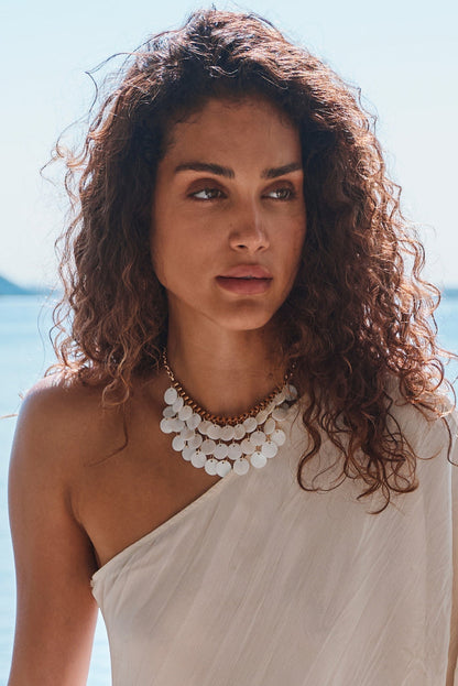 Layered white shell disc statement necklace on gold chain worn with a one-shoulder ivory dress by the sea.