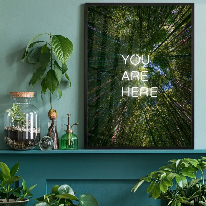 Framed You Are Here neon forest art print on archival paper, displayed on a teal shelf with indoor plants.