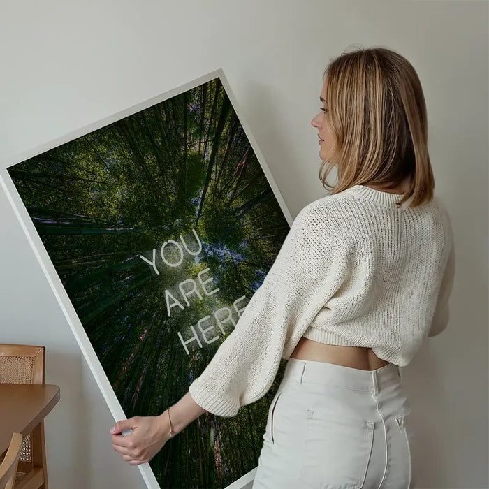 Woman holding a large You Are Here forest neon print in a white frame on archival art paper.