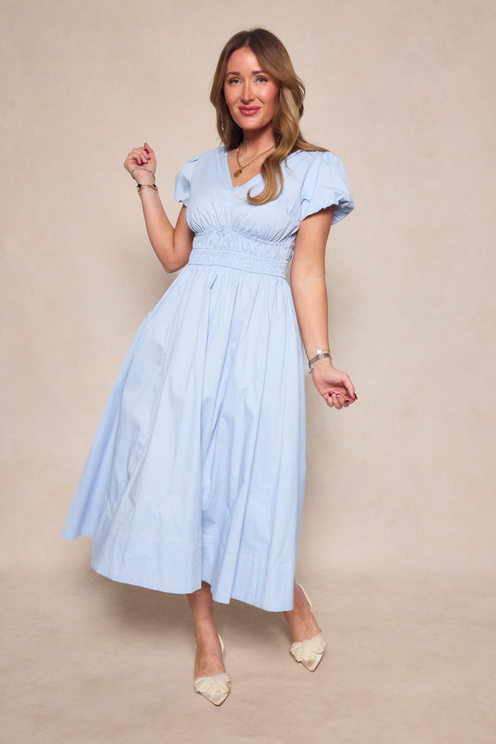 A woman poses against a neutral backdrop in the Ivy City Co Zella Dress in Sky Blue—a light blue, short-sleeve midi dress with a gathered waist and v-neckline—styled with bracelets, a necklace, and cream-colored shoes.
