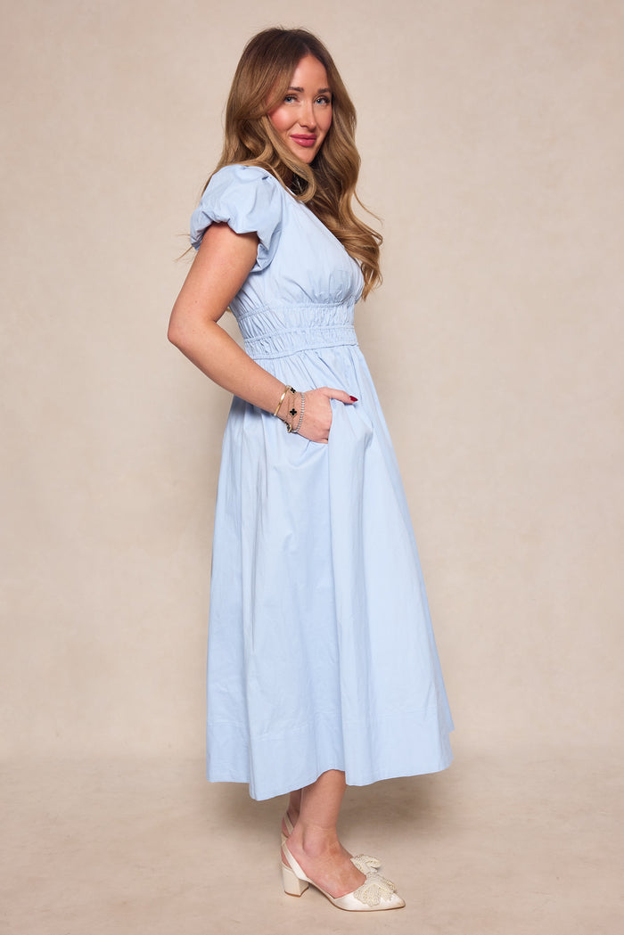 A woman with long wavy hair, wearing the Ivy City Co Zella Dress in Sky Blue with a smocked waist, stands sideways with one hand in her pocket and a slight smile. The ankle-length midi dress is paired with light-colored shoes.