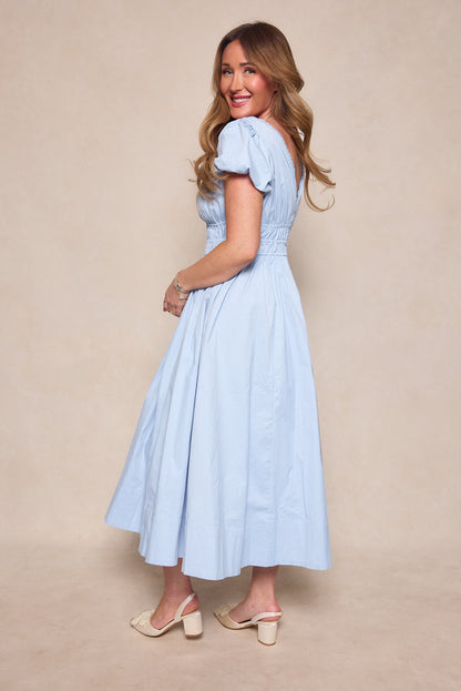 A woman with long, wavy hair smiles and stands sideways against a neutral background, wearing the Zella Dress in Sky Blue by Ivy City Co and cream heeled sandals.
