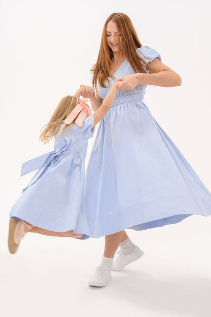 A woman and a young girl joyfully dance together in matching Ivy City Co Zella Dresses in Sky Blue, with the woman twirling the girl mid-air against a plain white background.