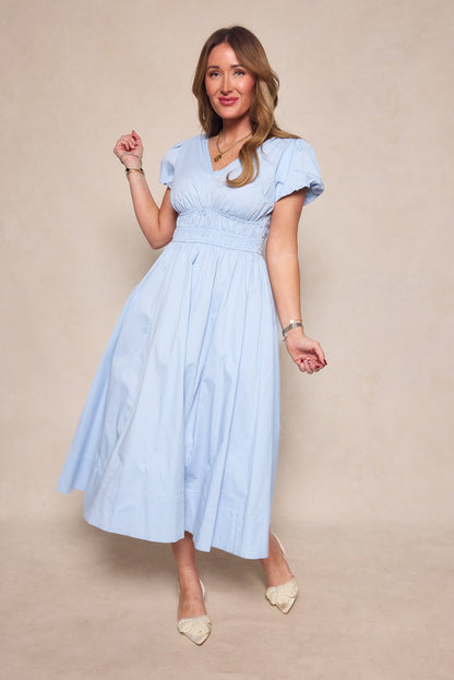 Wearing the Zella Dress in Sky Blue by Ivy City Co—a short-sleeve midi dress featuring a gathered waist—a woman stands on a beige background, smiling and posing with one hand raised. She completes her look with pointed beige shoes and gold jewelry.